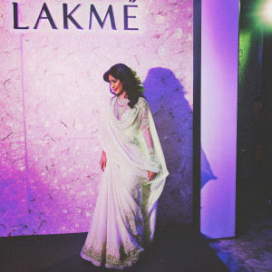 Chitrangada Singh at a high fashion event. Dressed in a stunning white saree with golden zari work, standing against a vibrant blue backdrop with the Lakmé logo.