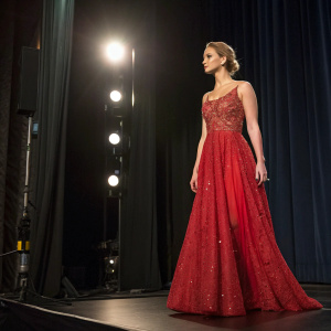 Jennifer Lawrence at the Gotham Awards. Wearing a flowing red dress with intricate beading. Standing alone on a black stage with spotlights.