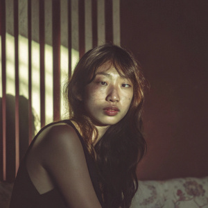 Photographic close-up of a young Asian woman with fair skin and long, wavy, dark hair. She's in her late teens with a neutral expression and a direct gaze. Wearing a sleeveless black top. Dramatic lighting highlights her face. Background is blurred, vertical blinds with a warm, reddish hue.