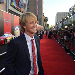 Owen Wilson, dressed in a navy suit with a crisp white shirt and a red striped tie, smiles warmly for photographers at the Cars 2 premiere. The background is a bustling red carpet lined with colorful, animated car characters.