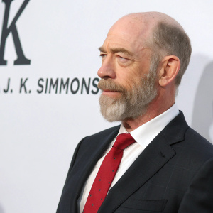 J.K. Simmons, mature man with a beard and balding head, wearing a tailored dark suit and a red tie. He is standing against a stark white backdrop with bold black text. Close-up shot.