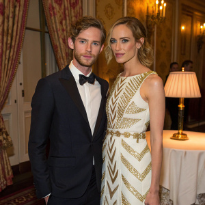 Luke Treadaway and Ruta Gedmintas at a gala event. Dark suit and bow tie for Luke, white and gold patterned dress for Ruta. Decorated with richly patterned curtains and elegant table with a lamp.