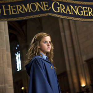 A young Emma Watson, as Hermione Granger, standing in front of a gold-embossed banner. She wears a blue Hogwarts robe and has a serious expression.