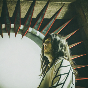 Close-up of Akira Geal, character with dark, flowing hair and striking black and white accents. Dramatic pose against an ominous, dark backdrop with sharp, angular geometric shapes in a mix of red, black, and gray hues.