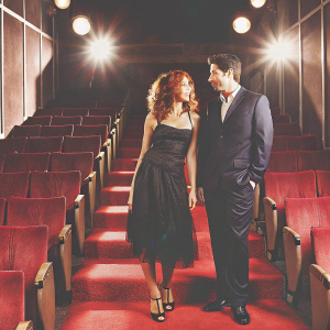 Zoe Buckman and David Schwimmer at Hollywood event. Zoe in elegant black dress. David in sharp navy suit. Dimly lit venue with spotlights