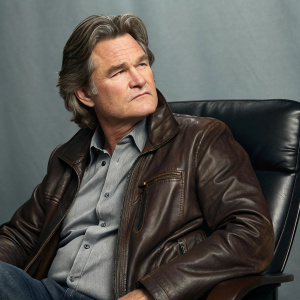 Kurt Russell, actor, intense expression. Sitting in an office chair, gray backdrop. Brown leather jacket, gray shirt. Professional photo style.
