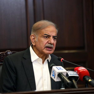 Shehbaz Sharif, Pakistani politician, addressing journalists. White shirt, dark suit, serious expression. Dark wood conference room backdrop.