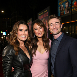 Brooke Shields, Lindsay Price, and Robert Buckley posing for a group photo at a Hollywood event. The background is dimly lit with various banners and posters of upcoming movies.