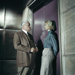 Charles Durning and Stephanie Faracy in a dramatic scene from the hit TV series 'Eye to Eye'. Charles is dressed in a classic brown suit and tie, while Stephanie wears a high-necked blue blouse and a patterned skirt. They share a serious look, set against a solid purple backdrop.