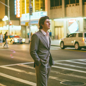 Garrett Hedlund, in a stylish charcoal grey suit, standing alone on a bustling city street at night. Neon lights cast colorful reflections around him.