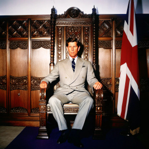 Prince Charles in a regal pose, sitting on an ornate throne. Grey tailored suit and patterned tie. Behind him, a wooden panelled wall adorned with intricate carvings and the English flag. High-definition photograph