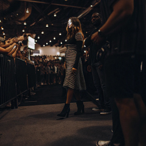 Hailee Steinfeld at Comic-Con, dressed in a chic black and white striped dress, paired with ankle boots. Crowded convention floor in the background.