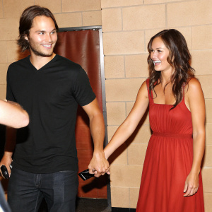 Taylor Kitsch and Minka Kelly at a fan meet and greet. Concrete wall backdrop. Both are dressed casually.