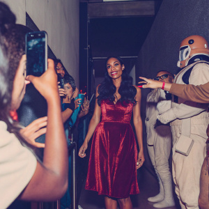 Rosario Dawson at the New York Comic Con. Dressed in a red velvet dress. Surrounded by excited fans and cosplayers.