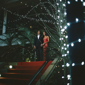 Carlo Conti and Tina Kunakey Di Vita at the Cannes Film Festival. Red carpet, glamorous evening attire, sparkling lights in background
