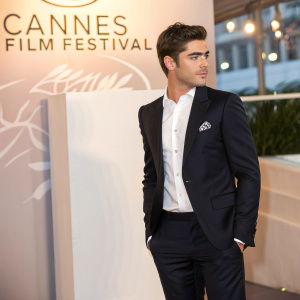Zac Efron at the Cannes Film Festival. Dapper in black suit, white shirt, and pocket square. Standing against backdrop with event logos and text.