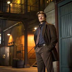 Nathan Fillion, standing confidently on the set of Castle. The background features a mix of industrial elements and warm, inviting lighting casting shadows on a textured wall.