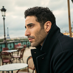 Close-up portrait of Miguel Angel Silvestre, actor. Dark curly hair, stubble, and a dark jacket. The setting is an outdoor cafe in Paris.