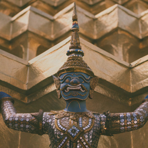 Close-up of a majestic guardian statue, featuring intricate blue, gold, and green mosaic patterns. The demon's arms are outstretched, conveying a powerful stance. Set against a backdrop adorned with golden Thai architecture and adorned with ornamental metallic accents and detailed patterns.