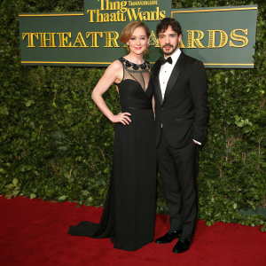 Hattie Morahan and Blake Ritson, at the Theatre Awards. Hattie in a sleek black gown with intricate neckline. Blake in a black tuxedo. Green hedge backdrop, red carpet, award banner