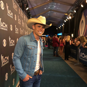 Dustin Lynch, white male, wearing cowboy hat and blue jeans, at the ACM Honors. Crowded backstage area with logos and banners.