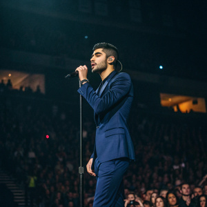 Zayn Malik at a concert venue. Dark blue suit, energetic pose, crowd blurred in background