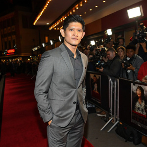 Iko Uwais, actor and martial artist, attending the premiere of his latest film. Wearing a tailored gray suit, standing confidently in front of a bustling crowd at the theater.