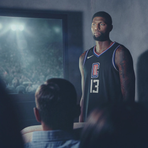 Paul George, NBA star, wearing black LA Clippers jersey with number 13. Serious look, athletic build with tattoos visible. Inside a packed arena, spotlighted, surrounded by cheering crowd.