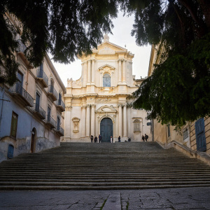The Baroque Church of Santa Chiara in Noto, Sicily, Italy. A stunning, creamy white facade adorned with intricate carvings and sculptures. Set against a cobblestone street lined with steps, bustling with sightseers and locals.