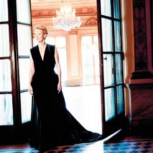 Tilda Swinton at the Met Gala, stunning in a sleek black velvet gown. Gown has deep neckline and floor-length skirt. She stands alone, the dark ballroom behind her.