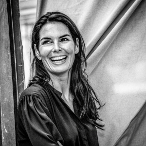 Close-up of actress Angie Harmon, smiling warmly. Long dark hair, black blouse, confident stance. Neutral toned, textured fabric backdrop.