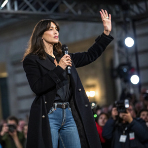 Monica Bellucci, Italian actress, with intense gaze. Wearing black coat and jeans. Speaking at an event, hands raised for emphasis. Crowd and flashing cameras in blurred background.