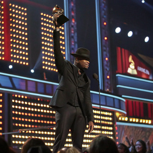 Ne-Yo accepting award at Grammy Awards. Dressed in a black suit, he raises his trophy amidst flashing lights and cheering crowd. Stage filled with LED screens displaying his nominations and accolades.