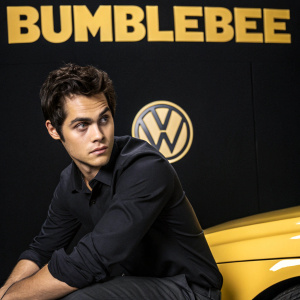 Dylan O'Brien in promotional shoot for 'Bumblebee'. Wears a dark shirt. Against black backdrop with 'Bumblebee' and VW logo in bold yellow letters. Intense stare