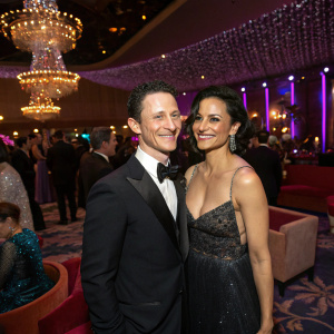 Jonathan Tucker and Zuleikha Robinson, two actors, at a glamorous Hollywood after-party. Lavish setting with sparkling chandeliers and celebrity guests.