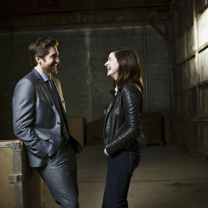 Jake Gyllenhaal and Anne Hathaway at a casual photo shoot. Both are smiling. Jake in a stylish suit, Anne in a leather jacket. Industrial setting with large boxes and bare walls.