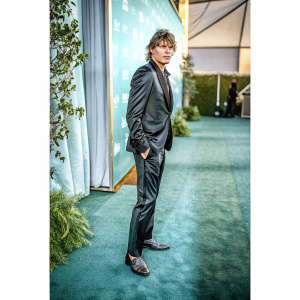 Jordan Barrett, sharp dressed in a sleek black suit, posing with a serious look at a gala. Dark blue backdrop with logo.