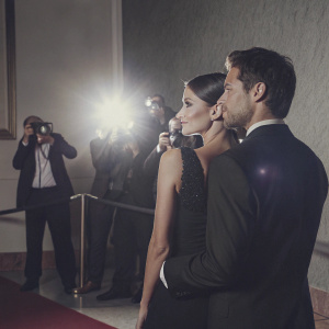 Sara Sampaio and Oliver Ripley at a glamorous awards ceremony. Sara wears a sophisticated black dress, Oliver in a black suit. Flashing lights from photographers capturing the moment