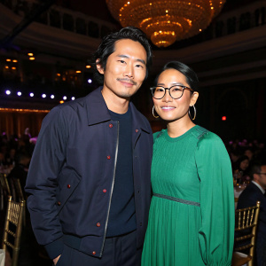 Steven Yeun and Ali Wong at an indoor award show. Wearing casual outfits. Steven in navy jacket, Ali in green dress. Backlit stage with shining lights