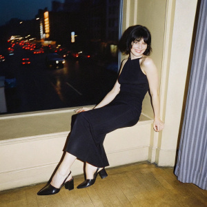 Kym Marsh, Caucasian woman, medium-length dark bob. Black sleeveless dress, black high heels. Posing confidently with a warm smile. Background: busy city street with neon lights.