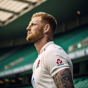 Ben Stokes, rugby player, intense stare. Dark tattoos, short beard. White rugby jersey. Indoor stadium, green seats in background.
