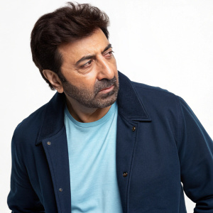Photograph of Sunny Deol, Indian actor. Focused expression, white background. Dark blue jacket over light blue t-shirt.