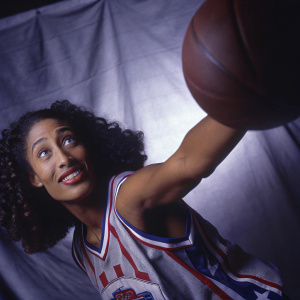 Skylar Diggins-Smith, basketball star, in a dynamic action pose. Wearing her team jersey with sponsors' logos. Curly dark hair framing her friendly face. Shot against a plain black background in a high-energy sports photo shoot.