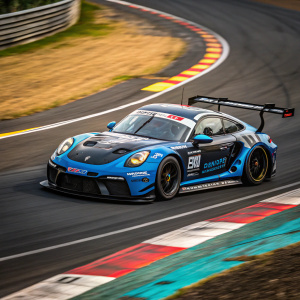 Porsche 911 RSR, blue and black livery, speeding on Nürburgring. Sponsor logos and decals visible. Colored track markings blurred in the background.