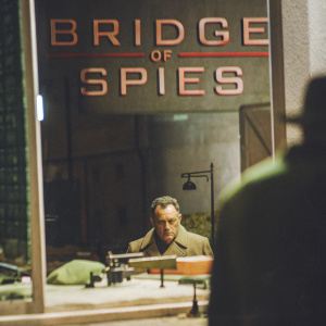 Film poster for 'Bridge of Spies'. Tom Hanks intense expression, framed against dimly lit Berlin backdrop. Cold-war era details, bridge prominently featured