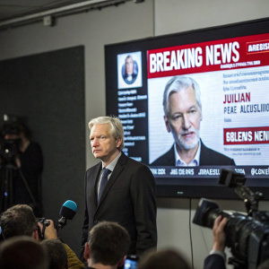 Julian Assange in a high-profile press conference. Standing in front of a large screen displaying breaking news headlines.