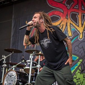 Randy Blythe, musician with dreadlocks, on stage at a punk rock concert. Dynamic pose with microphone in hand. T-shirt featuring a band logo. Drum set and graffiti-style backdrop with colorful spray paint designs.