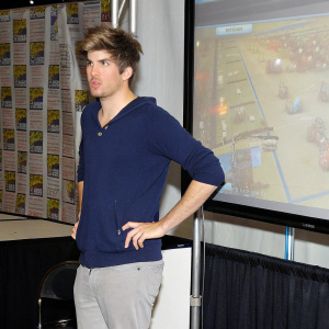 Joey Graceffa, appearing at a gaming convention. Wearing a casual blue hoodie. Large screen behind showcasing a gaming tournament