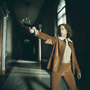 Tawny Cypress as Nez Rowan in action scene from 'The Blacklist'. Pointing gun in luxurious, old-world hallway. Dark, shadowy ambiance, sophisticated setting.