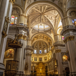 Interior of Granada Cathedral. Elaborate gold detailing, ornate paintings, multi-tiered architecture. Grand arch ceiling with intricate design. Stained glass windows bathe the space in soft, colorful light. Ornamental elements, paintings, and gold accents abound, creating a richly detailed environment.